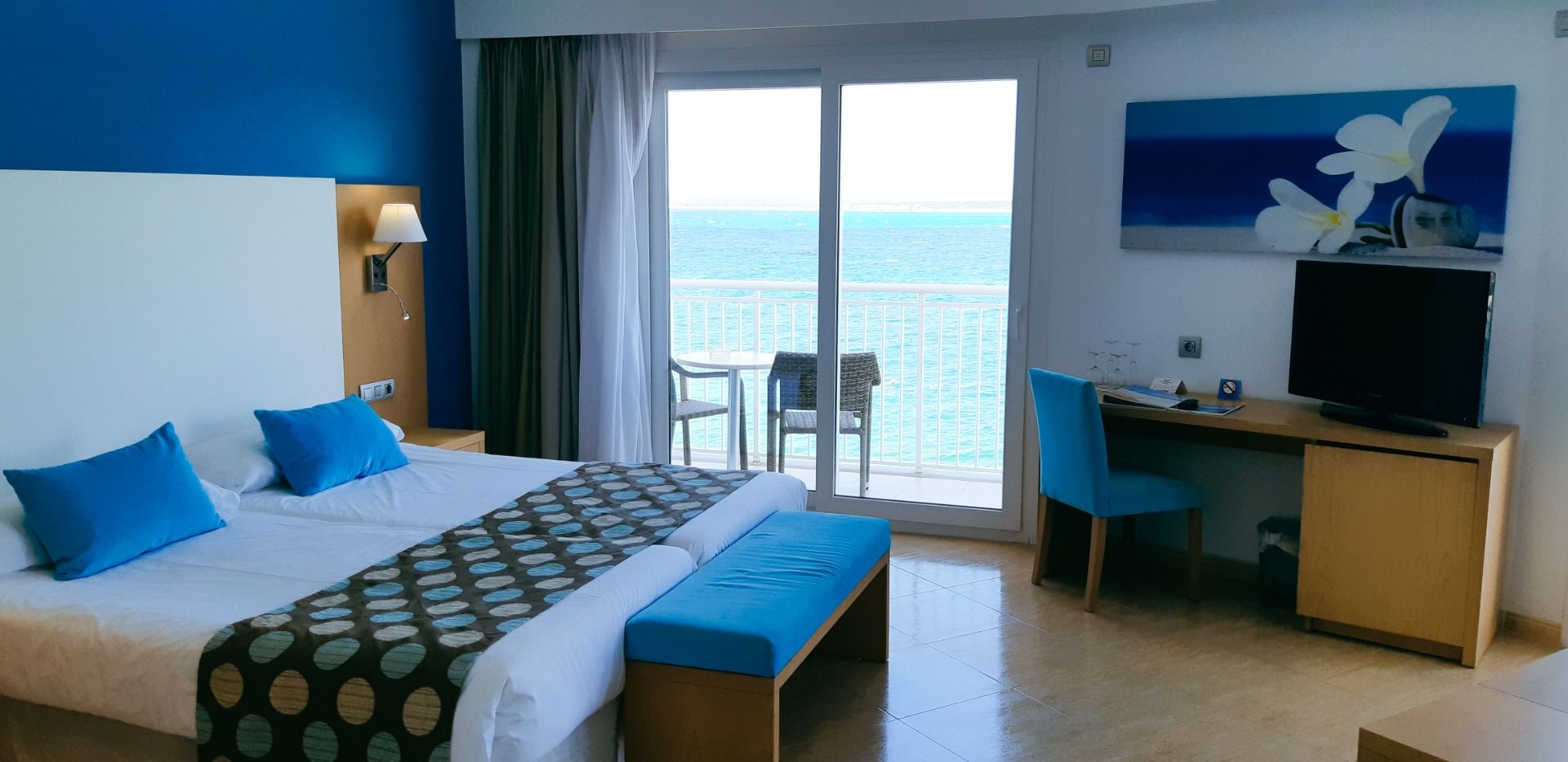 Helles Hotelzimmer mit großem Bett, blauer Einrichtung, Balkon mit Meerblick und einem kleinen Arbeitsbereich.