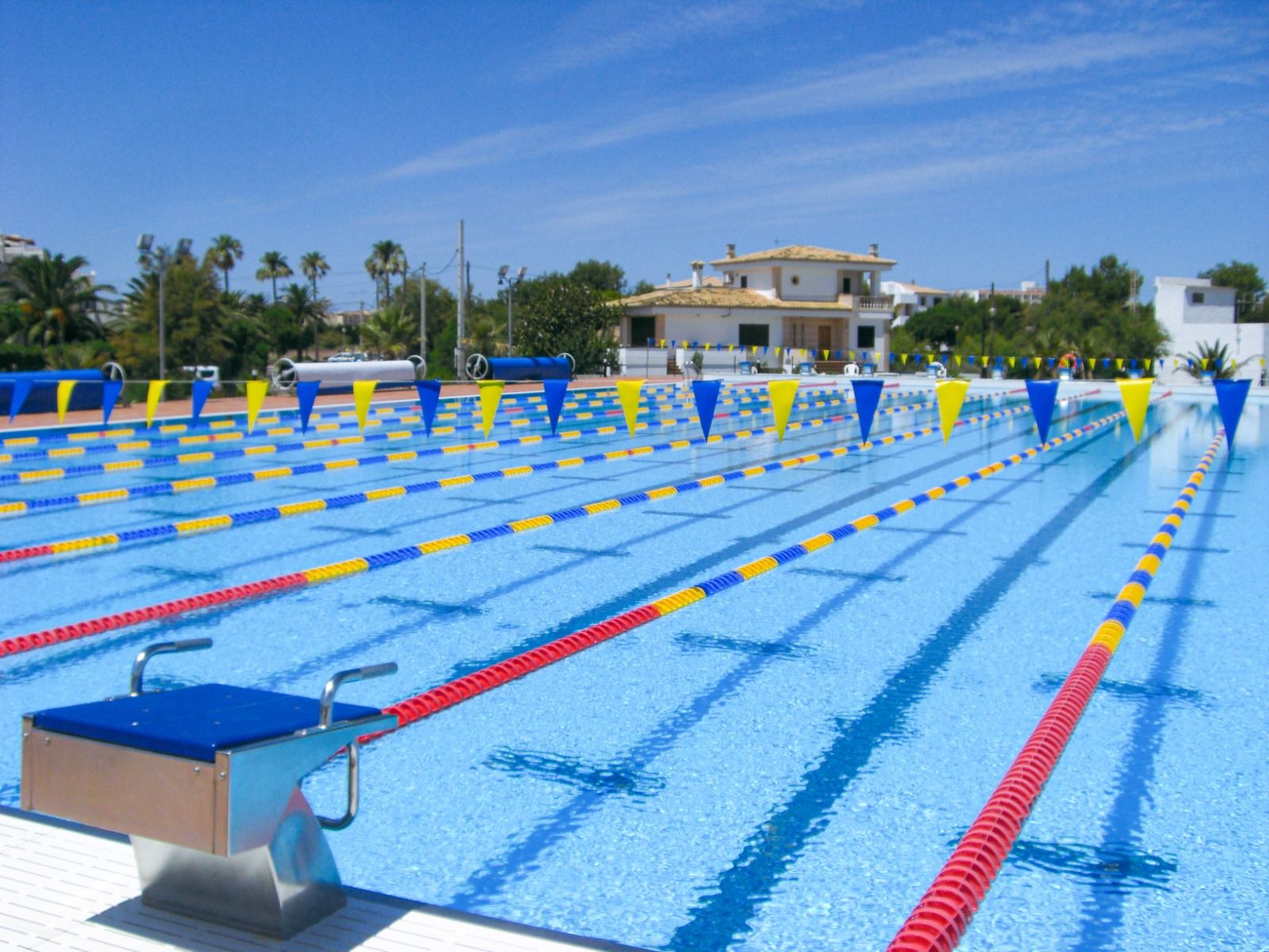Schwimmbecken mit klaren blauen Bahnen und Startblöcken, umgeben von Palmen und mediterraner Architektur.
