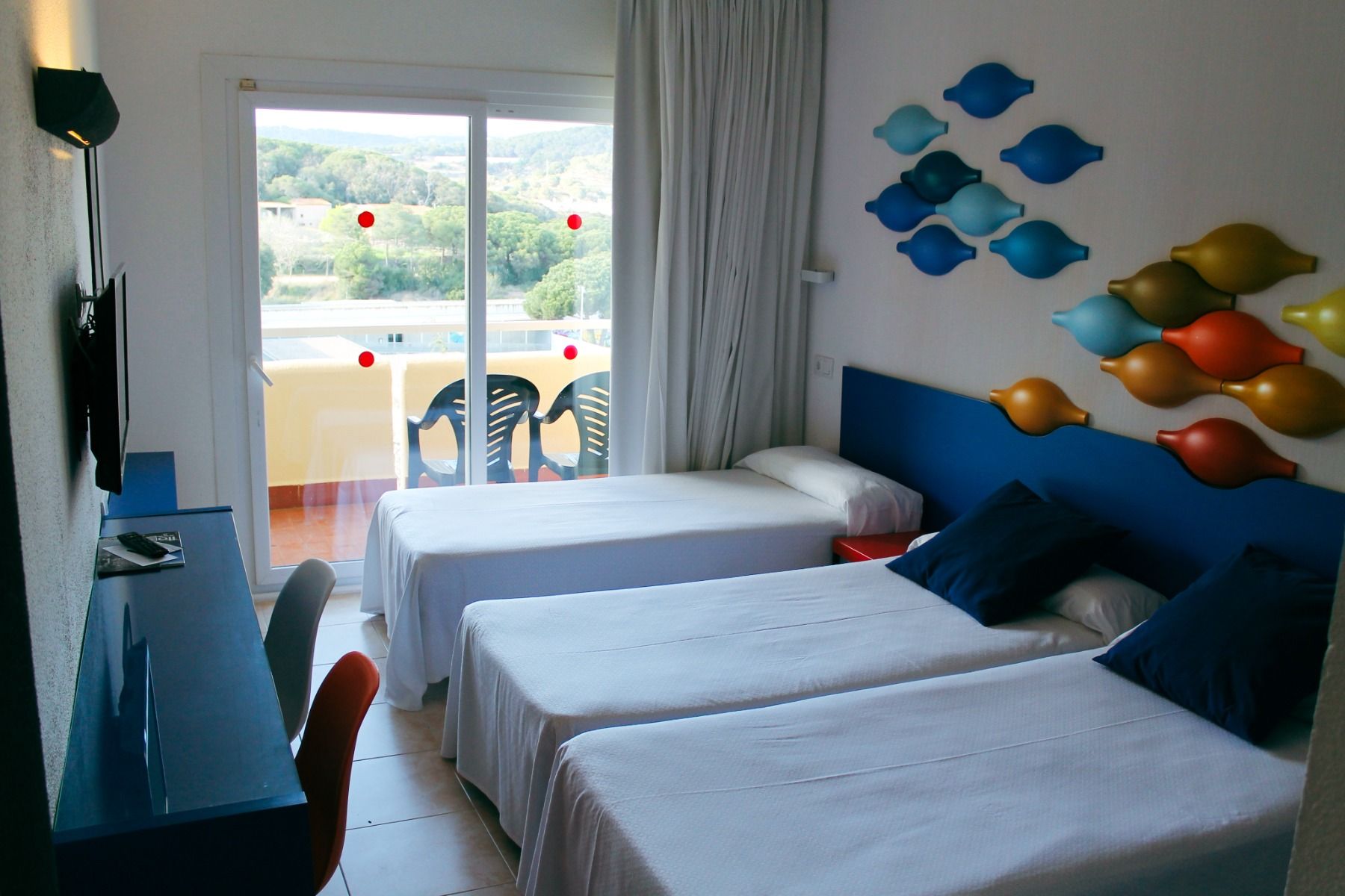 Hotelzimmer mit einem Einzelbett und einem Doppelbett, blauem Kopfteil und bunten Wanddekorationen, Zugang zu einem Balkon mit Blick ins Grüne.