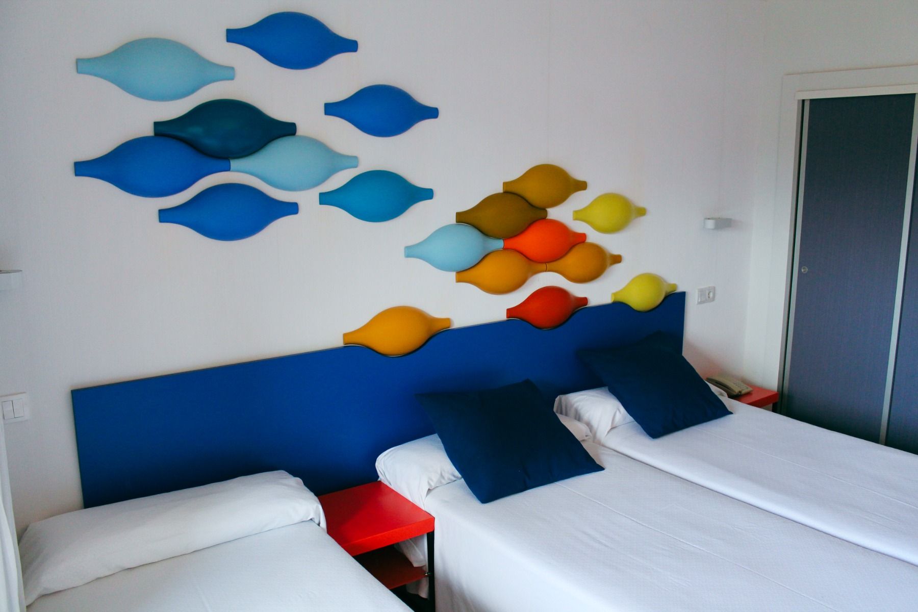 Modernes Hotelzimmer mit einem Einzelbett und einem Doppelbett, blauer Bettkopfteil und bunte Dekoration an der Wand.