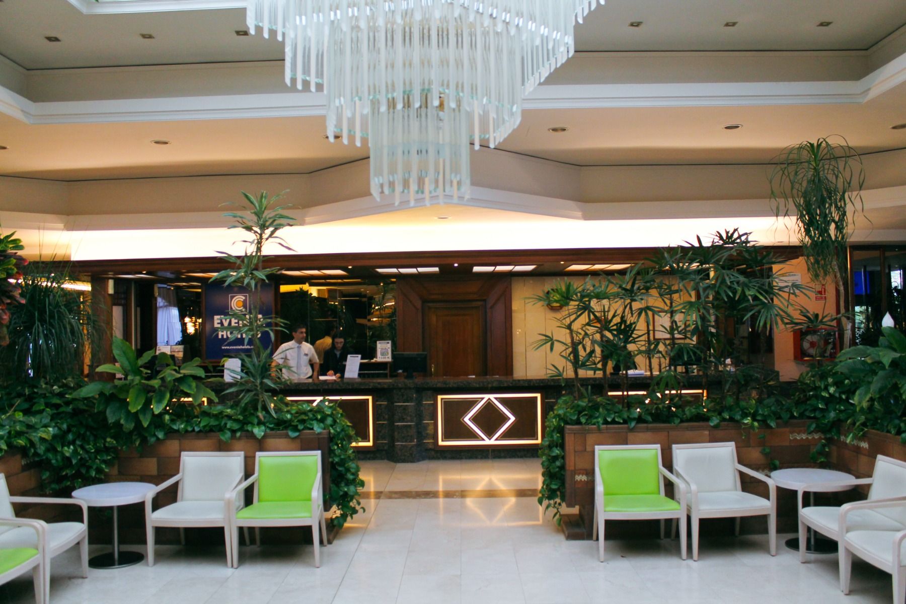 Empfangsbereich des Hotels mit vielen grünen Pflanzen, modernen Sitzgelegenheiten und einer Rezeption im Hintergrund.