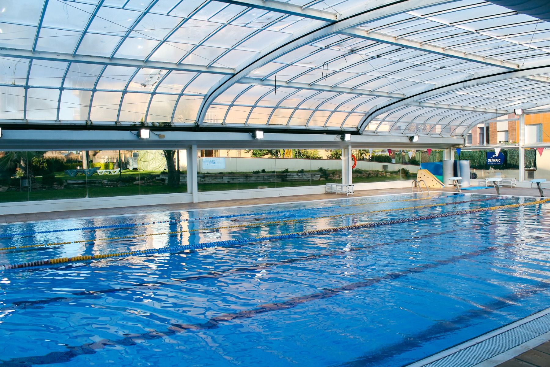 Überdachtes Schwimmbecken mit Glasdach, mehrere Schwimmbahnen und Sitzmöglichkeiten am Beckenrand.