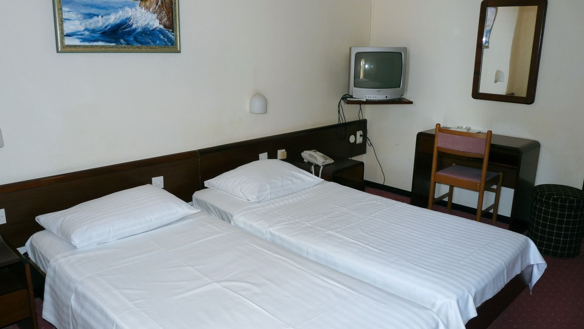 Ein schlichtes Hotelzimmer mit zwei Betten, einem kleinen Schreibtisch und einem Wandspiegel. Ein alter Fernseher steht auf einer Halterung