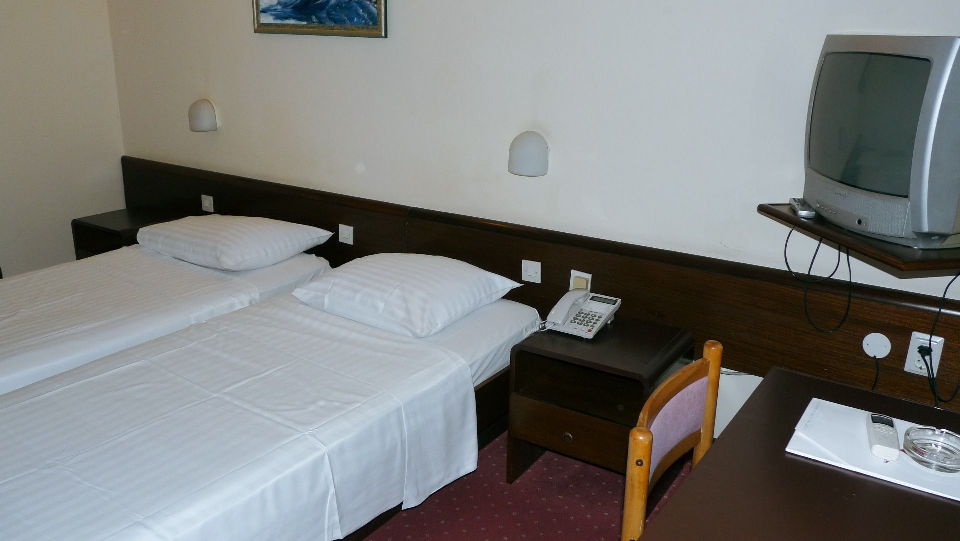 Ein einfach eingerichtetes Zimmer mit zwei Einzelbetten, Nachttischen und einer klassischen Lampe an der Wand. Im Hintergrund ist ein alter Röhrenfernseher auf einer Halterung sichtbar