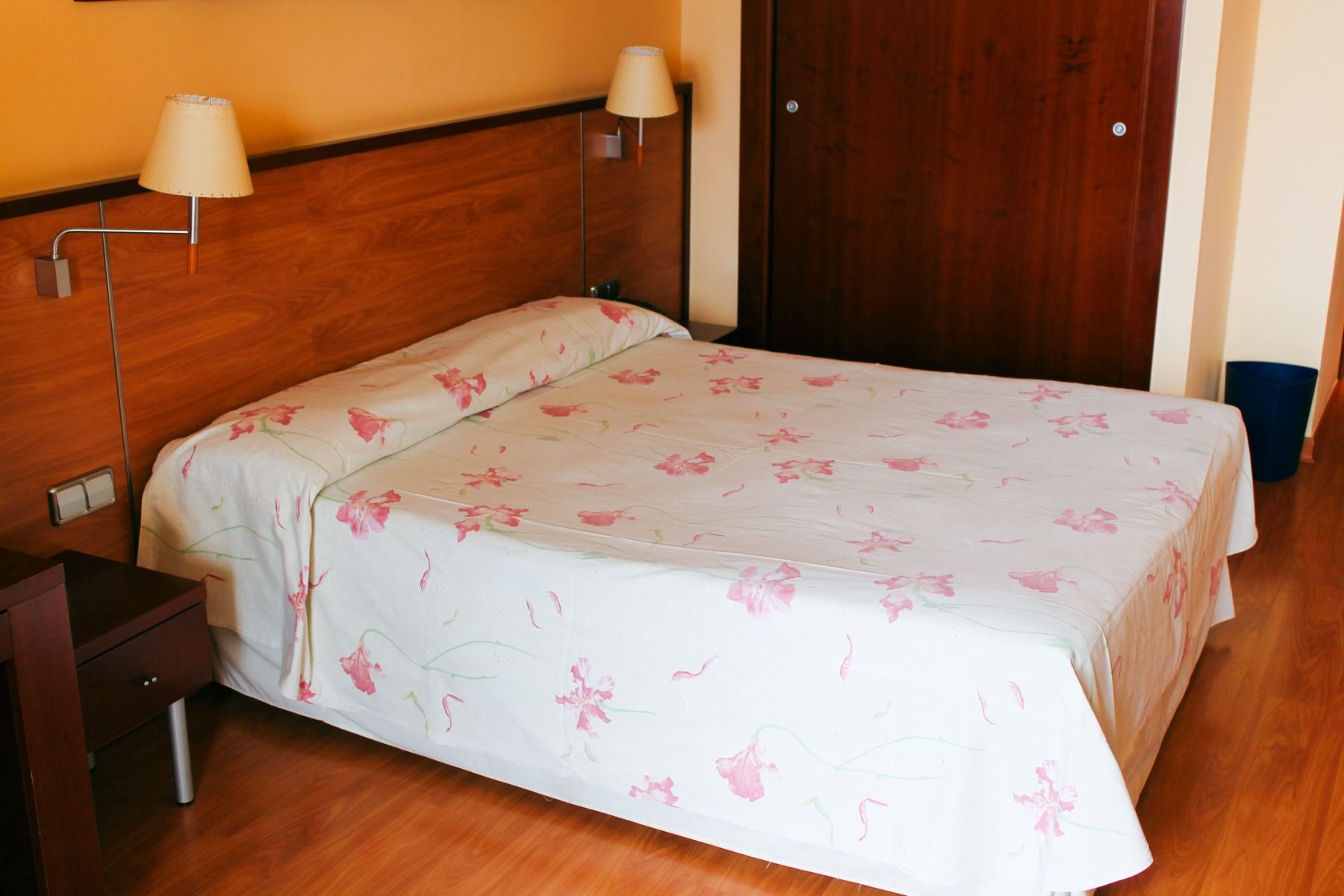 Hotelzimmer mit einem großen Bett, Holzoptik und rosa geblümter Bettdecke, gemütliche Einrichtung mit Nachttischen und Lampen.