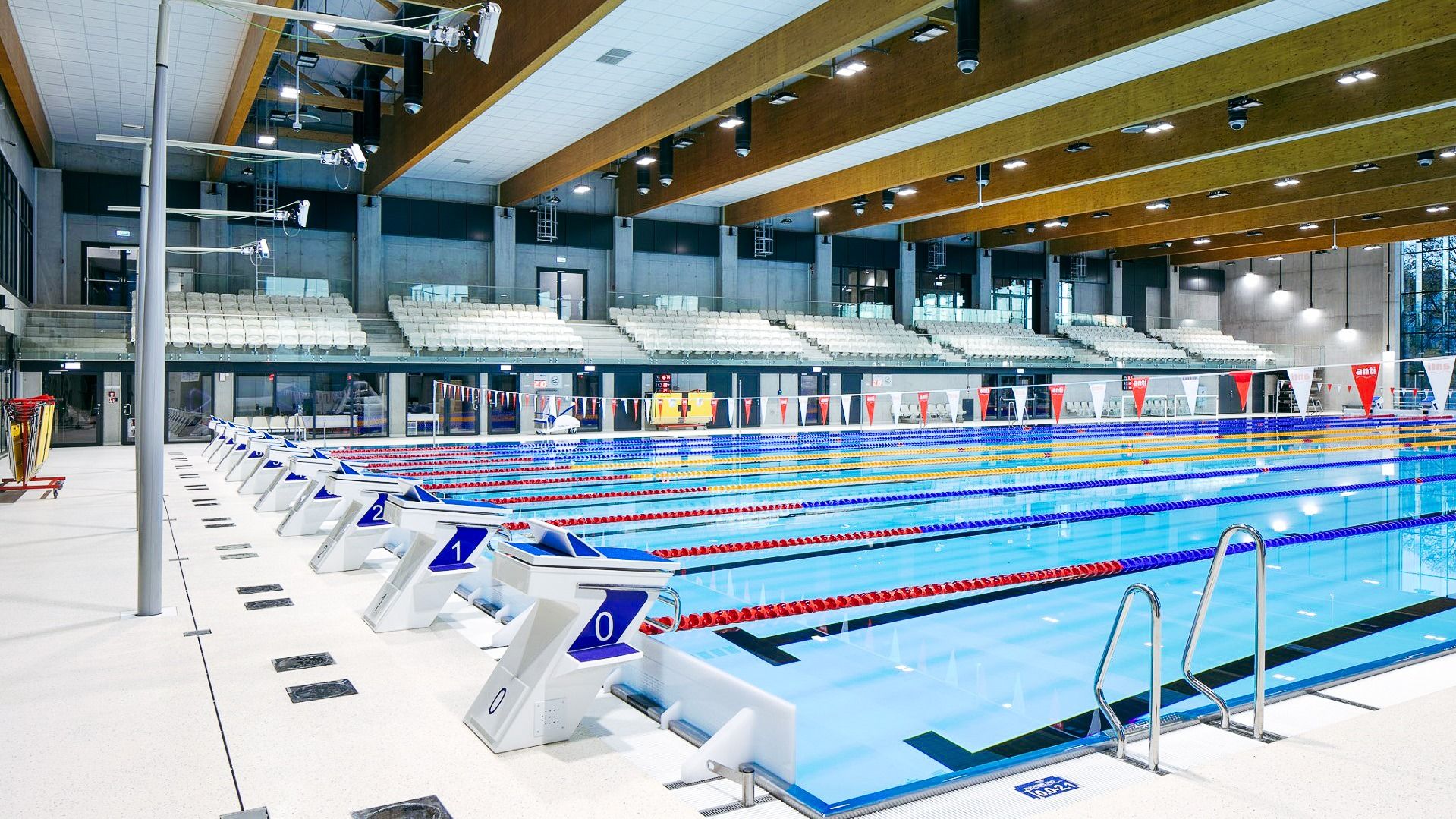 Innenansicht eines modernen Schwimmbads mit Wettkampfbecken, Startblöcken und Tribüne.