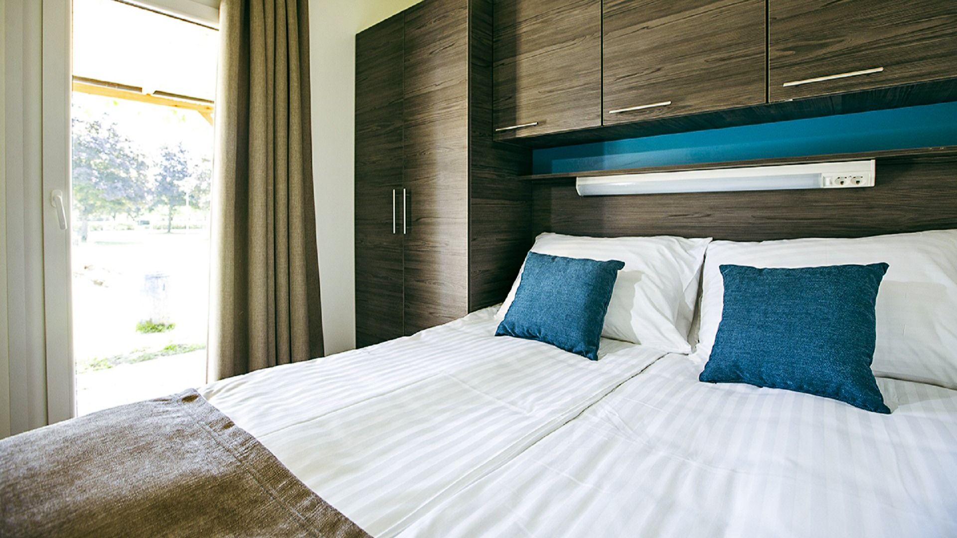 Gemütliches Schlafzimmer mit Doppelbett, blauen Kissen und moderner Holzvertäfelung im Mobile Homes Terme Ptuj.