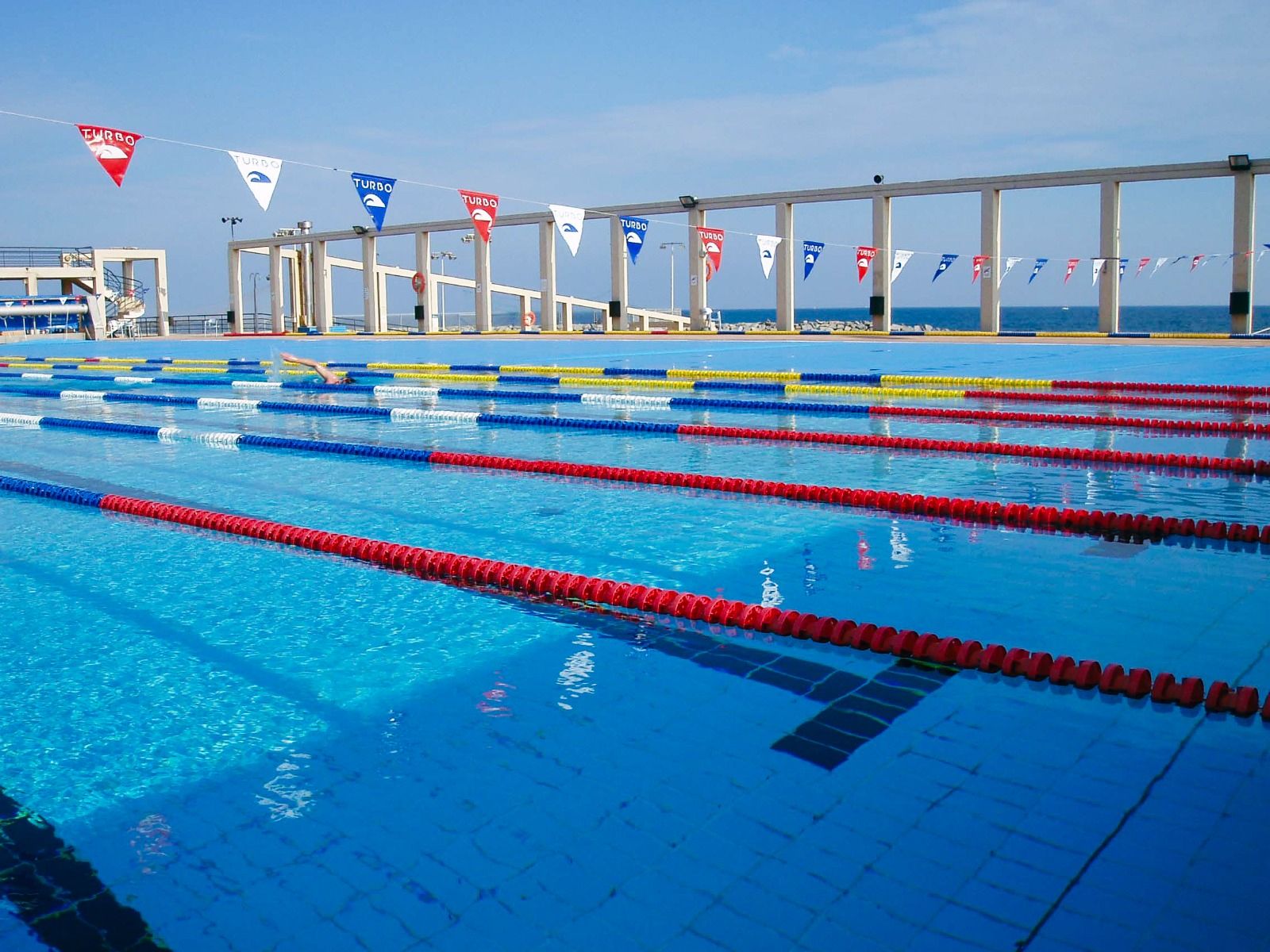 Ein großes Outdoor-Schwimmbecken mit abgetrennten Bahnen direkt am Meer, mit blauen, roten und gelben Markierungen und kleinen Fahnen zur Dekoration.