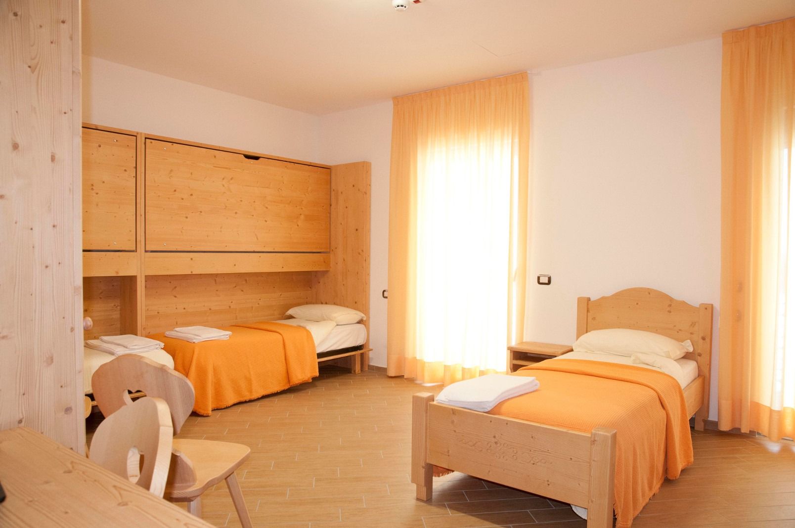 Großzügiges und freundliches Zimmer im Ostello Sportivo mit orangen Vorhängen, drei Betten mit orangen Decken, sowie Schrank, Tisch und Stühle aus Holz