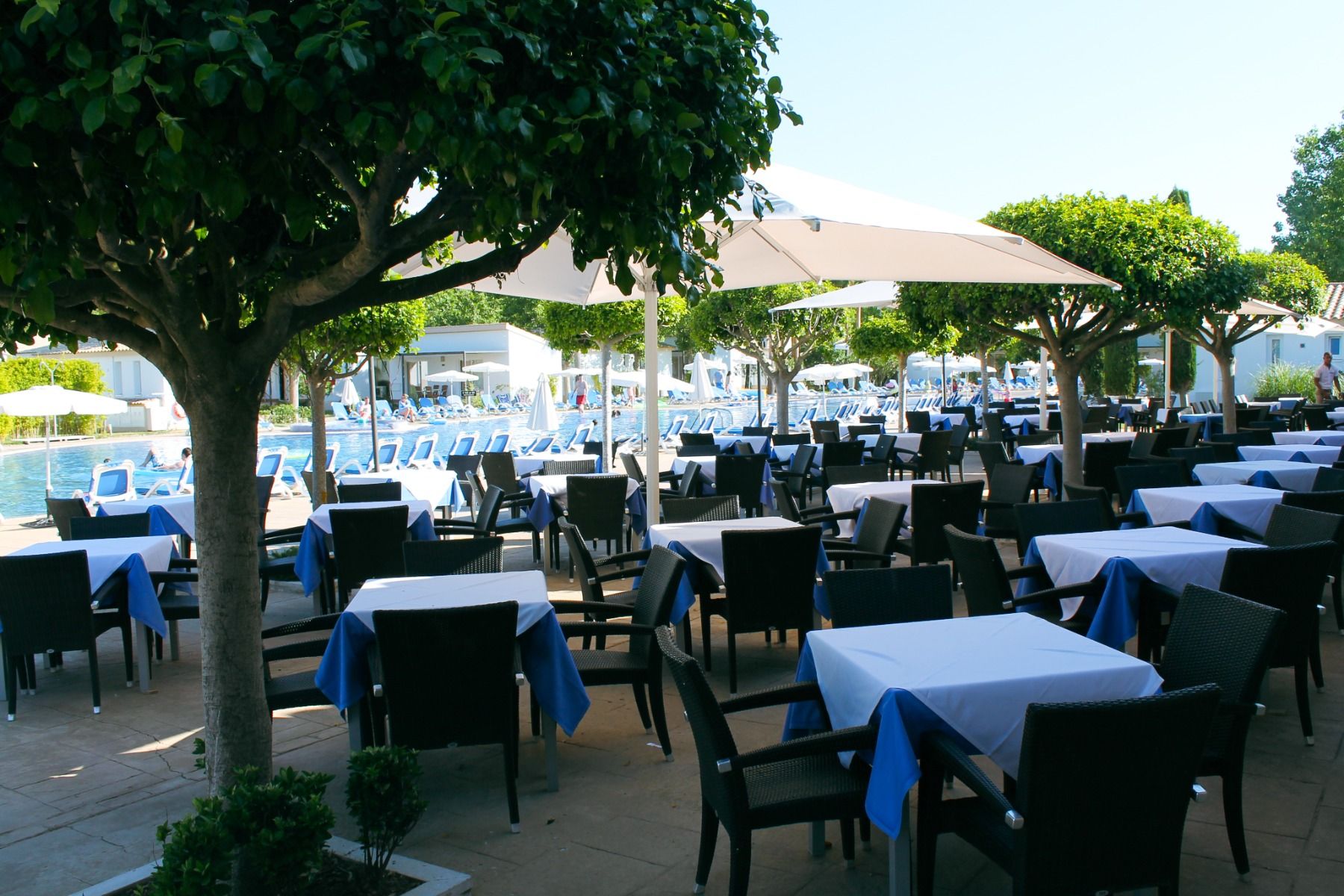 Außenbereich eines Restaurants mit Tischen und Stühlen, Sonnenschirmen, Bäumen und Pool im Hintergrund.