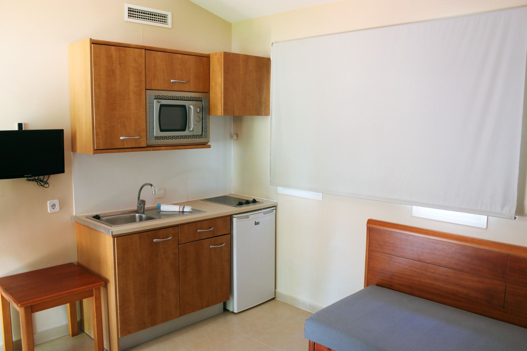 Kleine Hotelküche mit Holzschränken, eingebauter Mikrowelle, Kochplatte, Spüle und kleinem Kühlschrank, daneben ein graues Sofa.