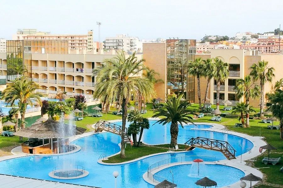 Weitläufiger Hotelgarten mit geschwungenem Pool, Palmen und einer Brücke, umgeben von Hotelgebäuden.