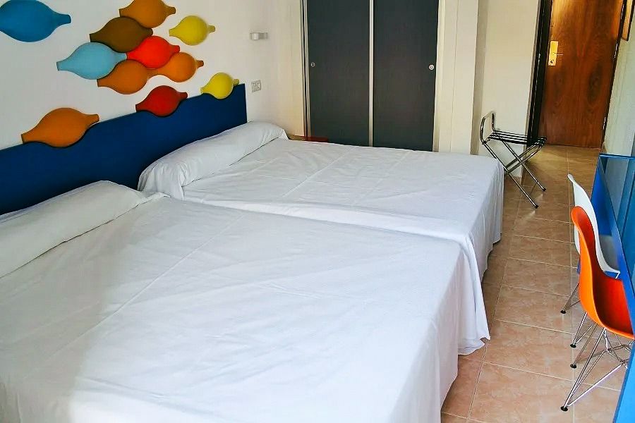 Helles Hotelzimmer mit zwei großen Betten, weißer Bettwäsche und einer bunten Wanddekoration, sowie einem Schreibtisch mit orangefarbenem Stuhl.