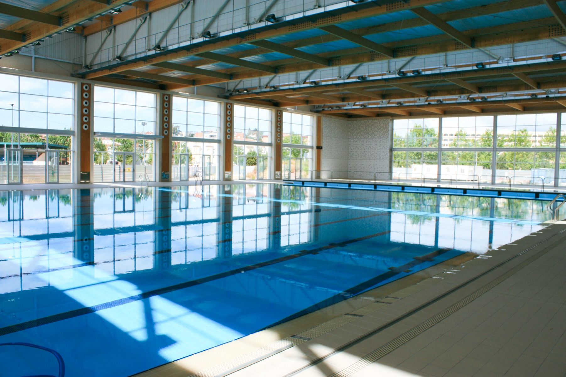 Moderne Schwimmhalle mit einem großen, hell erleuchteten Pool, hohen Glasfenstern und Holzbalkendecke.