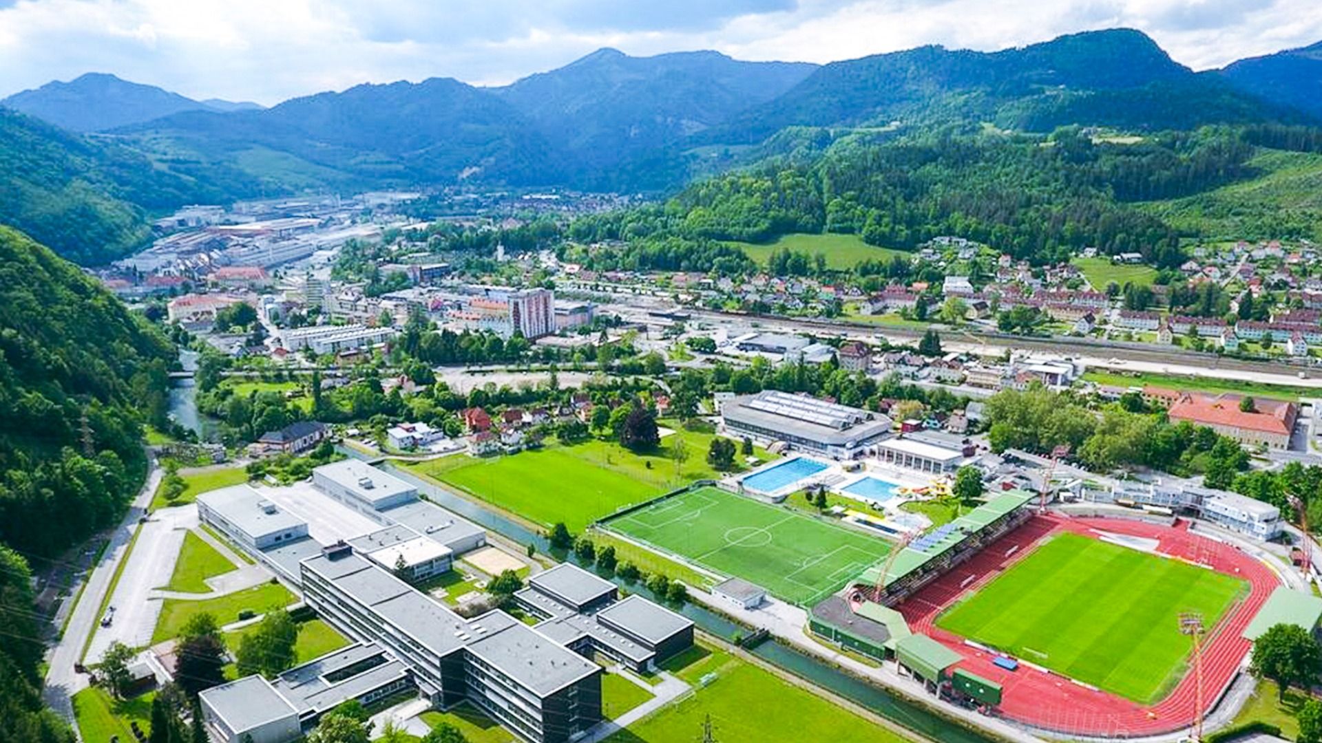 Luftaufnahme des Sportkomplexes Kapfenberg mit Schwimmbad, Fußballplatz, Leichtathletikstadion und umliegender Berglandschaft