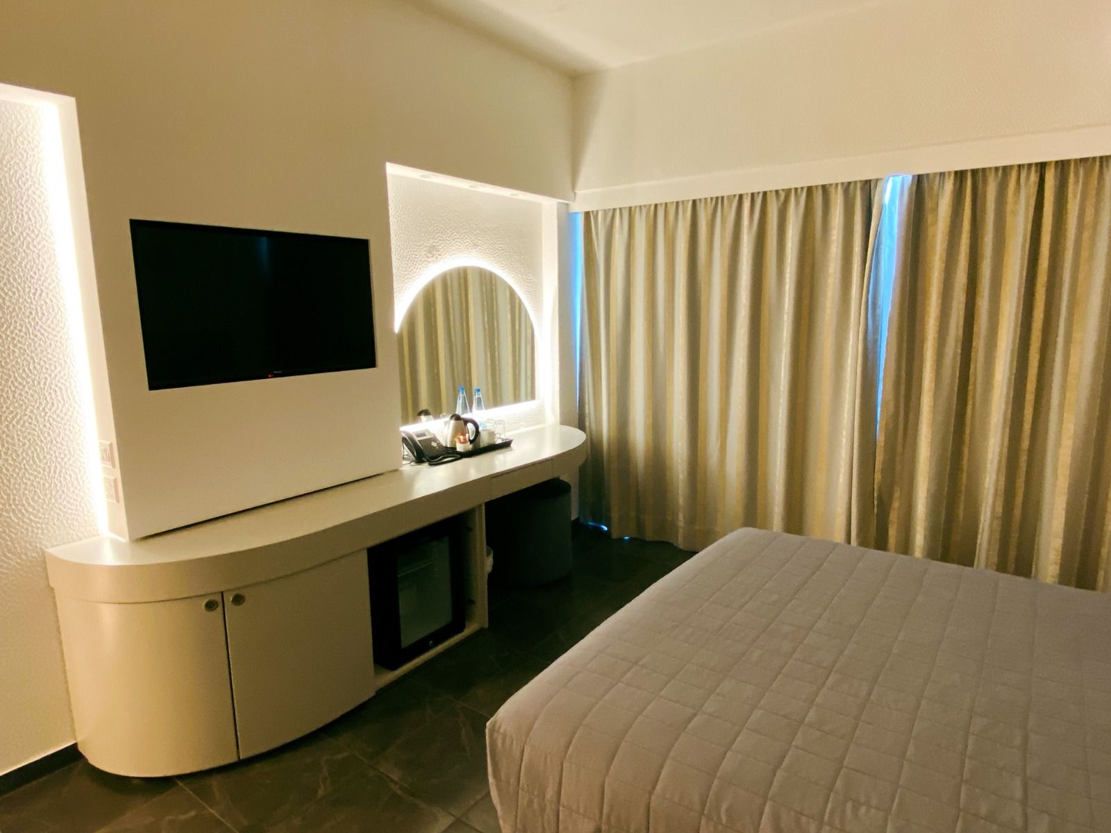 Hotelzimmer im Sveltos Hotel mit Bett, TV, Schreibtisch und Vorhängen, warm beleuchtet.