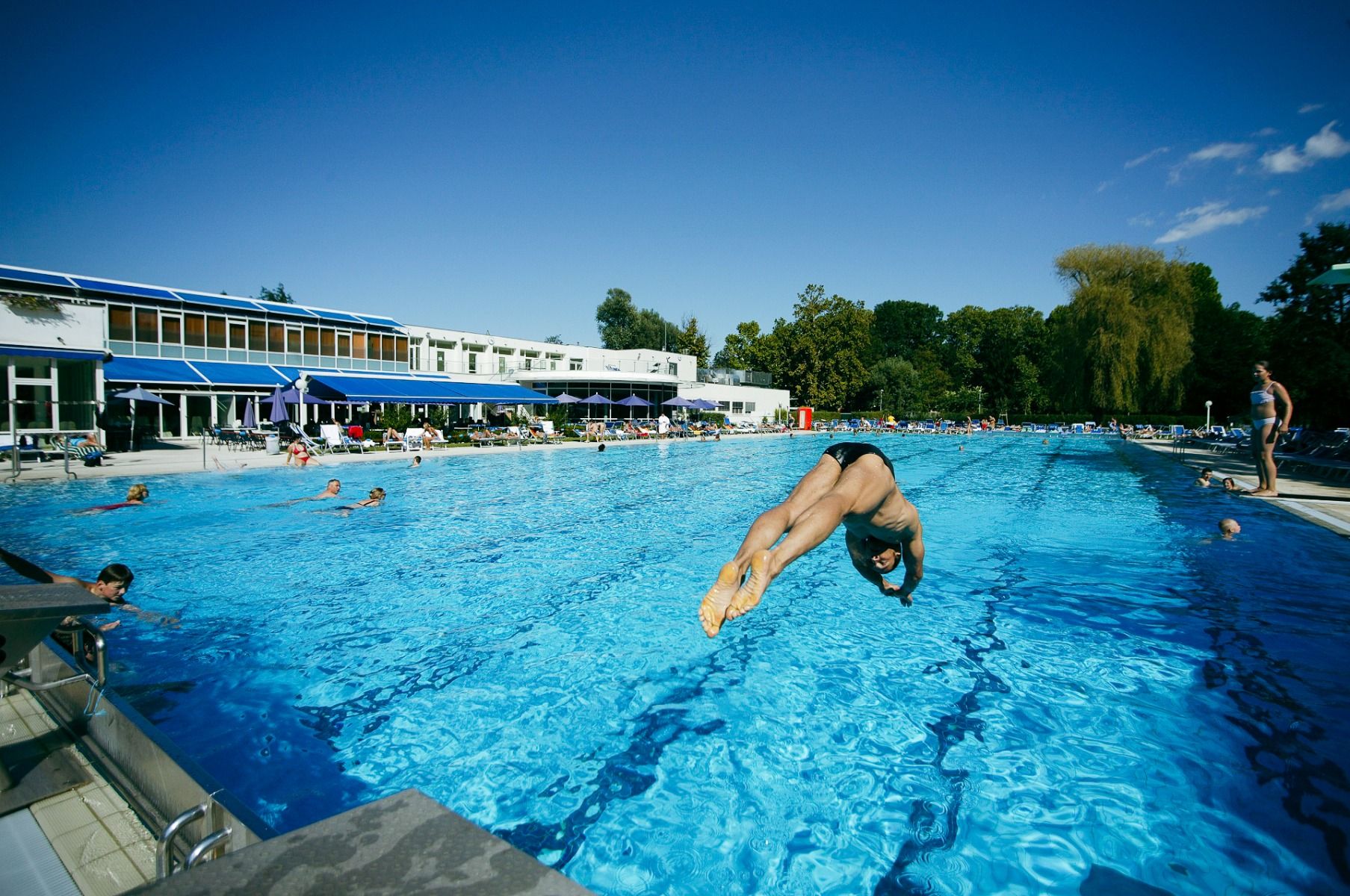 Großer Außenpool mit einer Person, die ins Wasser springt, umgeben von einer sonnigen Landschaft und klarem blauen Himmel.