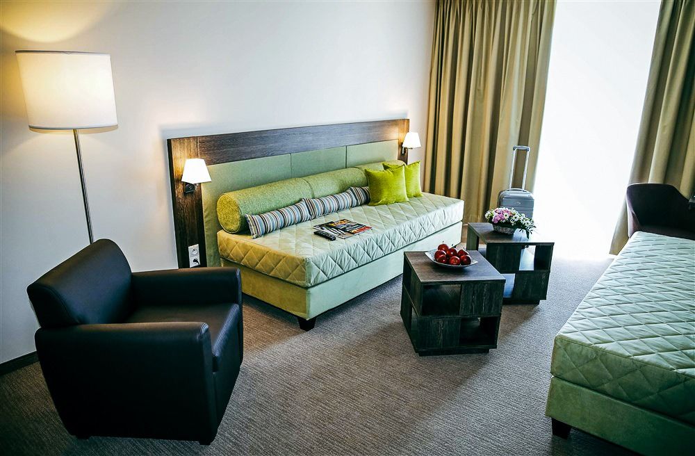 Komfortables Hotelzimmer mit grüner Couch, Beistelltisch mit Äpfeln, modernem Sessel und großem Fenster im X-Bionic Sphere Hotel.