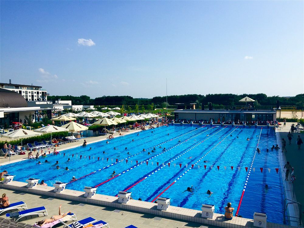 Outdoor-Schwimmbecken mit Schwimmern, Bahnenmarkierungen und Sonnenliegen unter Sonnenschirmen.