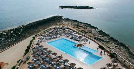 Ein modernes mehrstöckiges Hotel mit Balkonen an jedem Stockwerk, darunter ein Poolbereich mit Sonnenschirmen und entspannten Gästen bei klarem Himmel.