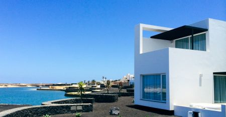 Moderne weiße Hotelvilla mit Panoramafenstern und überdachter Terrasse, umgeben von schwarzem Sand und Blick auf eine ruhige Lagune.