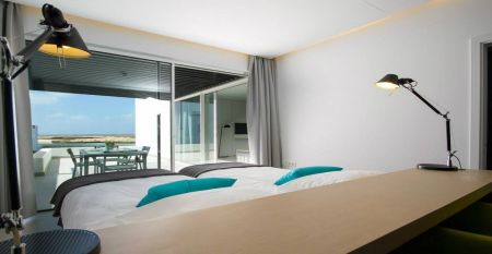Moderne weiße Hotelvilla mit Panoramafenstern und überdachter Terrasse, umgeben von schwarzem Sand und Blick auf eine ruhige Lagune.