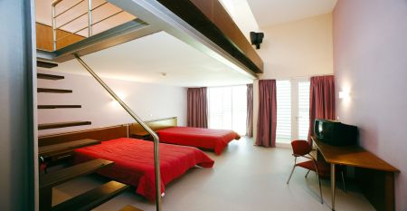 Helles Hotelzimmer mit zwei Betten mit einer roten Tagesdecke. Zudem eine Treppe, die zu einer zweiten Ebene im Zimmer führt.