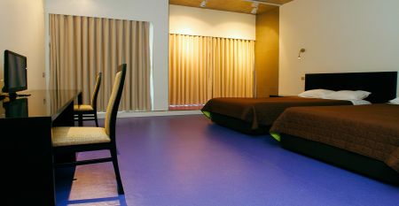 Helles Hotelzimmer mit zwei Betten mit einer roten Tagesdecke. Zudem eine Treppe, die zu einer zweiten Ebene im Zimmer führt.