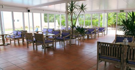 Hotelterrasse mit Pool, umgeben von Liegestühlen und Tischen mit Stühlen, angrenzend an ein gelbliches Gebäude mit Balkonen und großen Glasfenstern.
