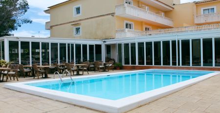 Hotelterrasse mit Pool, umgeben von Liegestühlen und Tischen mit Stühlen, angrenzend an ein gelbliches Gebäude mit Balkonen und großen Glasfenstern.