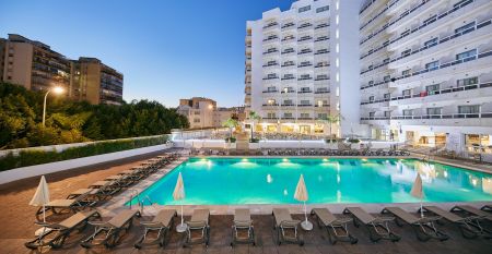 Hotelanlage bei Dämmerung mit beleuchtetem Pool, Sonnenliegen und mehrstöckigem weißen Gebäude.