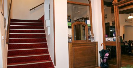 Der Empfangsbereich des Hotels Koruna, mit einer hölzernen Rezeption und einer roten Teppichtreppe, die nach oben führt.