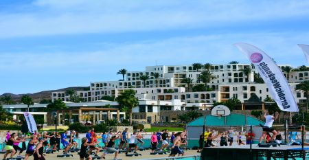 Outdoor-Fitnesskurs mit einer Gruppe von Menschen, die gemeinsam auf Steppern trainieren, vor der Kulisse einer weißen, terrassenförmigen Hotelanlage.