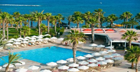 Große Poollandschaft umgeben von Palmen und Sonnenliegen vor dem Louis Phaethon Beach Hotel mit der weißen Hotelanlage im Hintergrund.