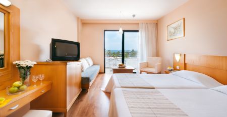 Große Poollandschaft umgeben von Palmen und Sonnenliegen vor dem Louis Phaethon Beach Hotel mit der weißen Hotelanlage im Hintergrund.