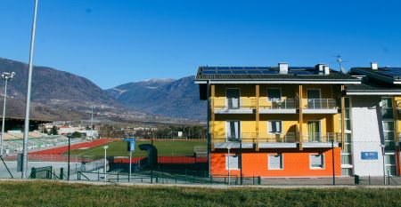 Außenansicht des Ostello Sportivo mit organge-gelb gestrichener Fassade, dahinter ein Fußballplatz und Berge