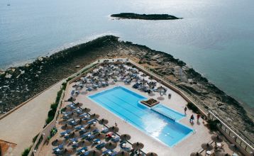 Ein modernes mehrstöckiges Hotel mit Balkonen an jedem Stockwerk, darunter ein Poolbereich mit Sonnenschirmen und entspannten Gästen bei klarem Himmel.