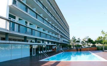 Außenansicht des Hotels mit Balkonen und einem großzügigen Poolbereich mit Liegestühlen und Holzboden.