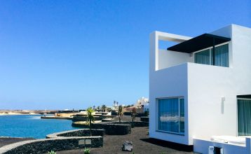 Moderne weiße Hotelvilla mit Panoramafenstern und überdachter Terrasse, umgeben von schwarzem Sand und Blick auf eine ruhige Lagune.