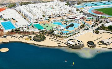 Moderne weiße Hotelvilla mit Panoramafenstern und überdachter Terrasse, umgeben von schwarzem Sand und Blick auf eine ruhige Lagune.