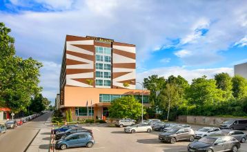 Ein modernes Hotelgebäude mit auffälligen geometrischen Mustern in Braun und Weiß, umgeben von grünen Bäumen und einem großen Parkplatz im Vordergrund.