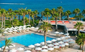 Große Poollandschaft umgeben von Palmen und Sonnenliegen vor dem Louis Phaethon Beach Hotel mit der weißen Hotelanlage im Hintergrund.
