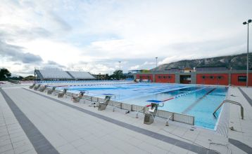 Fußweg auf der Anlage mit bunten Unterkunftshäusern umrahmt von Bäumen im Sport Camp in Loutraki, Korinthia